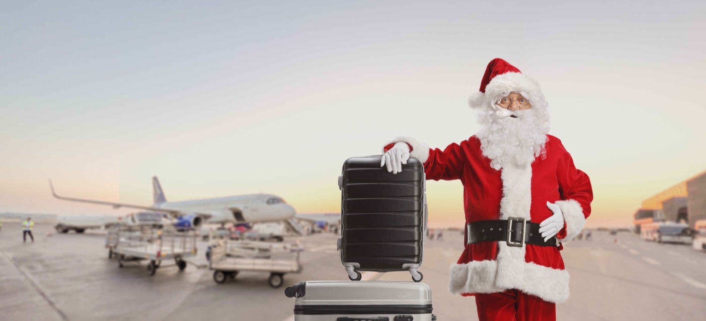 Santa Claus at an airport with a suitcase near an airplane.